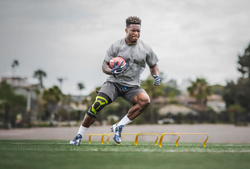 A college football player performing footwork drills at practice while holding a football. Athlete wearing a knee brace designed to treat iliotibial band syndrome (ITBS), highlighting its use during athletic activity.